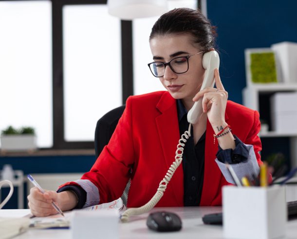 Businesswoman taking notes on clipboard sitting at desk in corporate office, while taking with adviser about financial expertise. Busy manager having a conversation about deadline.